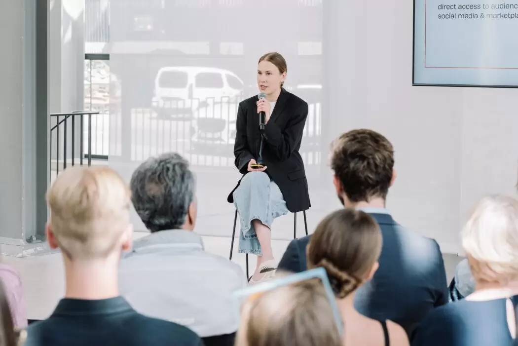 Evelina Gorbacova, Head of Digital Development at FOLLOW.ART, speaks into a microphone during her presentation at Riga Contemporary Art Fair 2025. She is seated in front of a screen and an attentive audience. Photo by Dasha Trofimova.