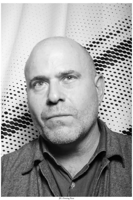 Black-and-white portrait of Marc Spiegler, a bald man with a close-cropped beard, wearing a button-up shirt and jacket. He stands against a graphic dotted backdrop from JR’s Printing Press photobooth installation.