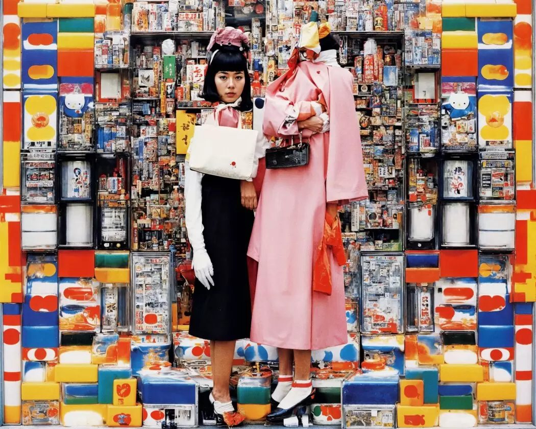 Two women stand against a dense, colorful backdrop of Japanese vending machines and packaging in The Altar of Bonnō (2024) by Emi Kusano. One figure wears a black skirt, white gloves, and carries a white bag, while the other in a bright pink coat appears partially distorted—reflecting Kusano’s AI-generated exploration of human desire and the Buddhist concept of Bonnō.