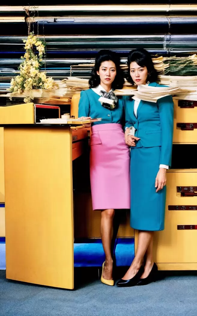 Two women in brightly colored retro office uniforms pose against a backdrop of stacked files in Office Ladies by Emi Kusano. Generated with AI, the scene highlights gender roles and stereotypes in corporate culture, blending surreal visuals with commentary on modern office life.