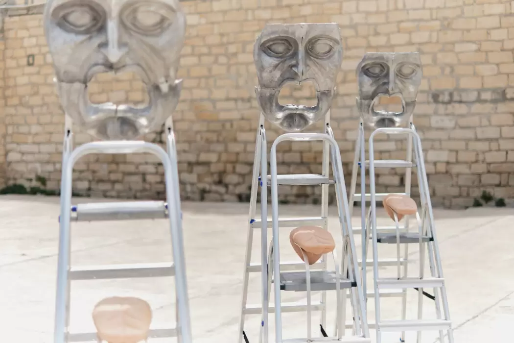 Three sculptural installations by Panos Profitis featuring metallic expressive masks mounted on ladders with padded seats, set against a stone wall in an outdoor space.