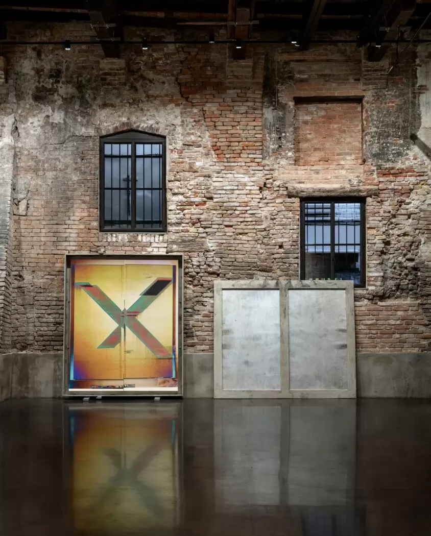 Wade Guyton, Untitled (2021), a large inkjet-on-linen painting with a bold “X” motif in orange and neon hues, displayed alongside Untitled (2024), a cast aluminium panel. Installed against the exposed brick walls of AMA VENEZIA.