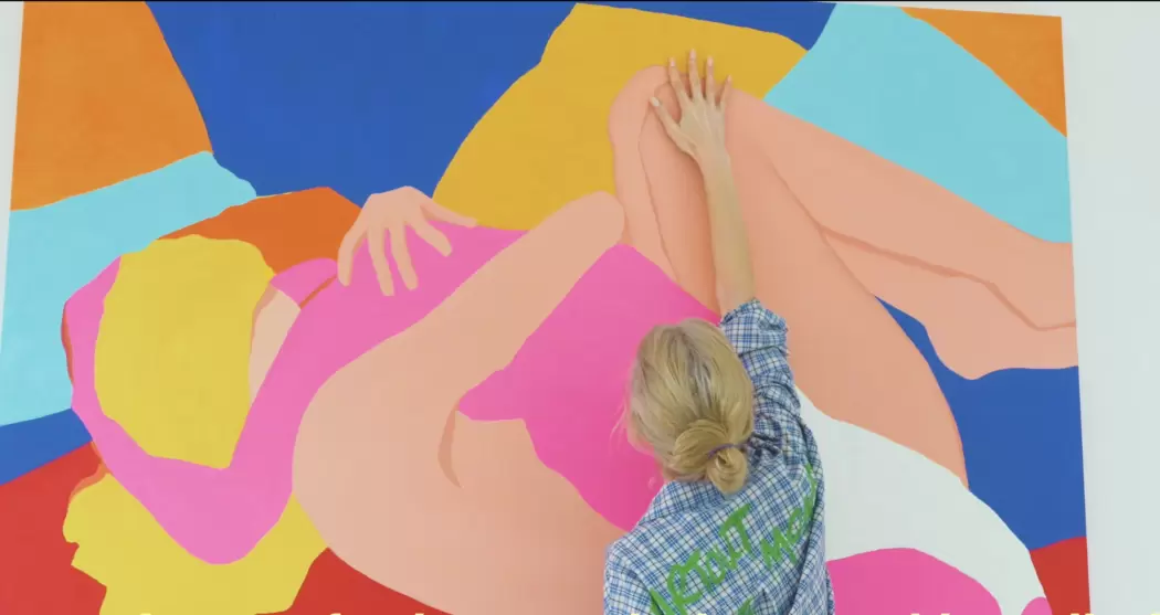 Anna Voda, seen from behind in a checkered shirt, reaches up to paint a vibrant, large-scale artwork of reclining figures in bright pink, orange, blue, and yellow tones inside her studio.