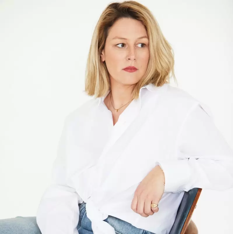 Portrait of Pauline Foessel, founder of 100 Collectors, sitting in a chair wearing a white shirt and light blue jeans against a white background.