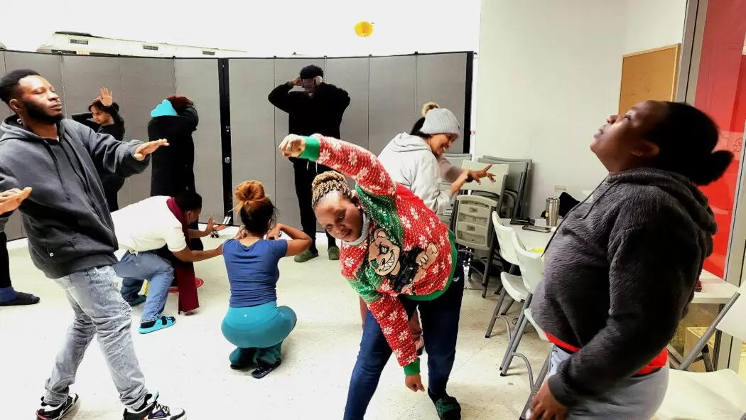 A group of adults in a classroom-like space participating in a drama therapy exercise, each striking different expressive poses. Some lean, stretch, or gesture with their arms, creating a lively and dynamic scene. The room has chairs stacked to the side and a neutral background divider.