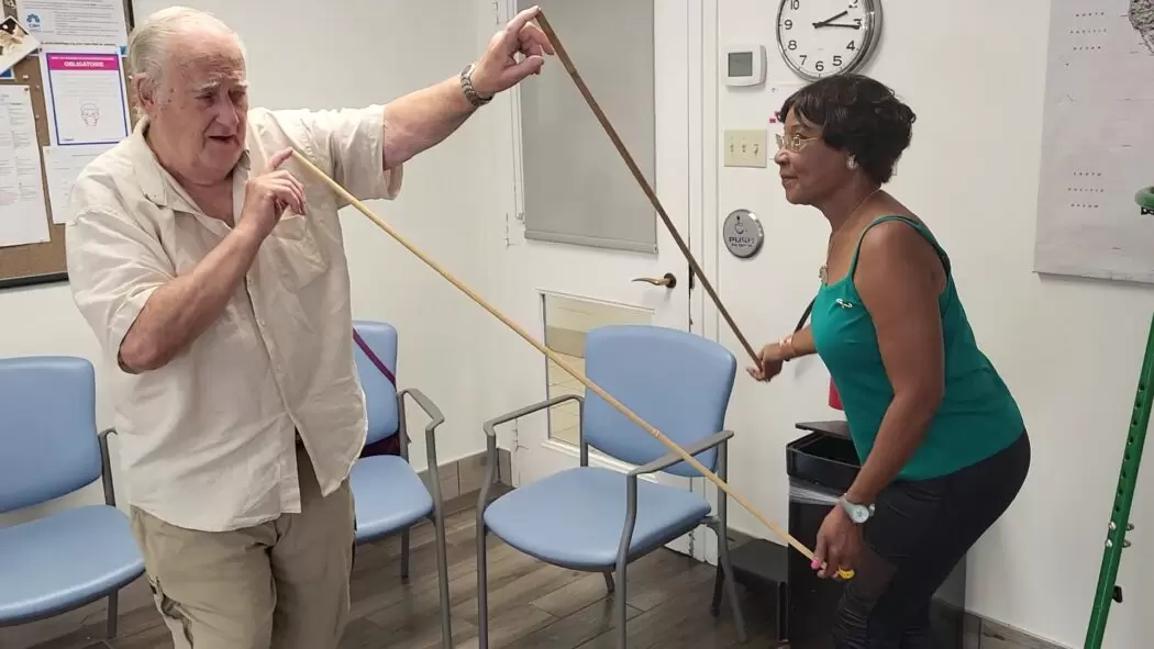 Two older adults participating in a drama therapy activity, each holding a long bamboo stick and moving in coordination. They are in a bright room with chairs arranged around them, creating a calm and supportive environment for senior-focused sessions.