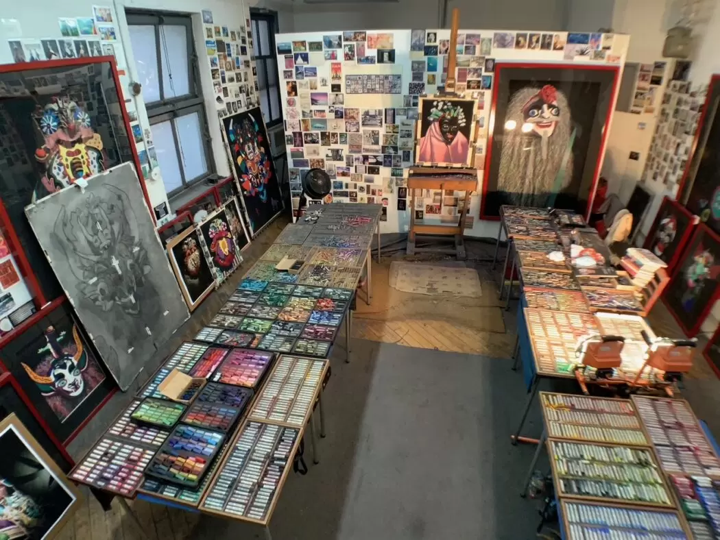 View of Barbara Rachko’s art studio featuring tables covered with hundreds of pastel sticks organized by color, large framed paintings depicting colorful mask-like figures, and walls lined with reference photos and sketches.