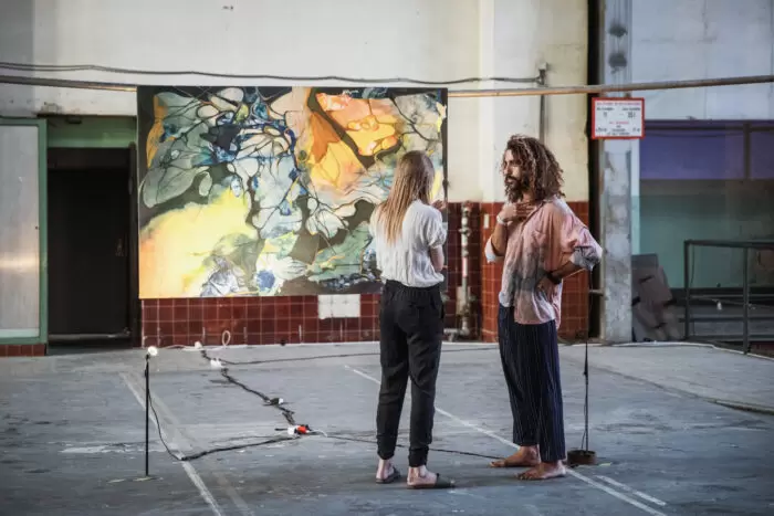 Two people stand barefoot in a spacious, industrial interior, facing each other in conversation. Behind them hangs a large abstract painting with organic, cell-like forms in blue, yellow, and earthy tones, lit by floor-mounted lights, suggesting an informal exhibition or workshop environment.