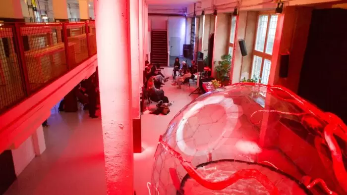 Interior view of a large, red-lit exhibition space with an audience seated for a talk, seen from above. In the foreground, a transparent, dome-like interactive sculpture glows red, with tubular elements inside, evoking an abstract eye and inviting sensory engagement.