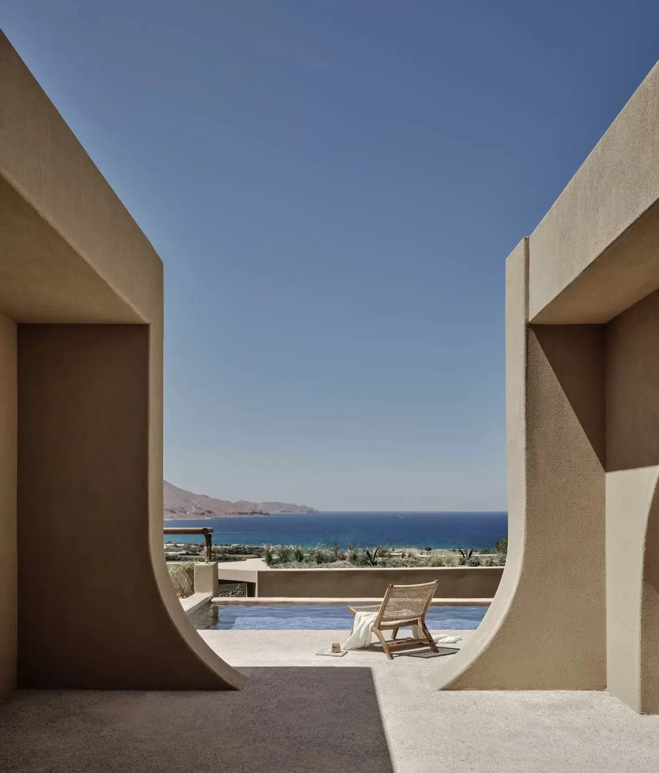 View through curved beige architectural walls at Tella Thera Hotel in Crete, framing a pool and lounge chair overlooking the sea, with natural light and minimalist design creating a calm, restorative atmosphere.