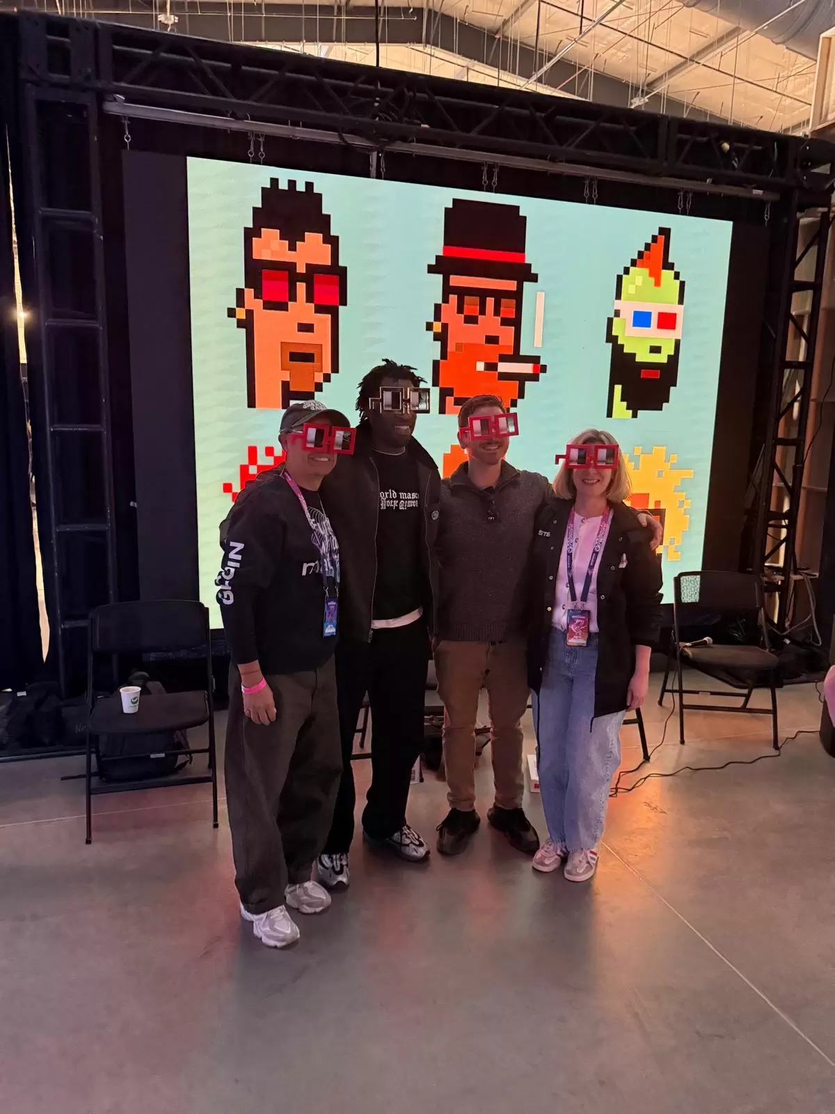 Group of visitors wearing pixel-style glasses posing in front of a large CryptoPunks display screen at the ETHDenver Art Gallery.