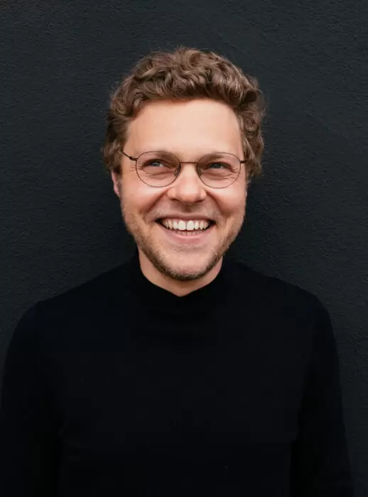 Portrait of Oleg Stavitsky smiling against a dark textured wall, wearing round glasses and a black turtleneck, looking slightly off-camera.