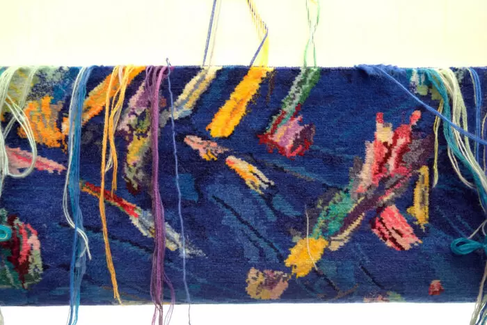 Close-up of a partially woven carpet titled Winter, with loose multicolored threads hanging over a deep blue background, translating Medina’s abstract imagery into textile form.