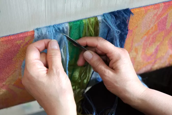 Close-up of hands weaving a colorful carpet on a loom, using blue, green, and warm-toned threads, as part of Medina’s work Flowers for My Mother.
